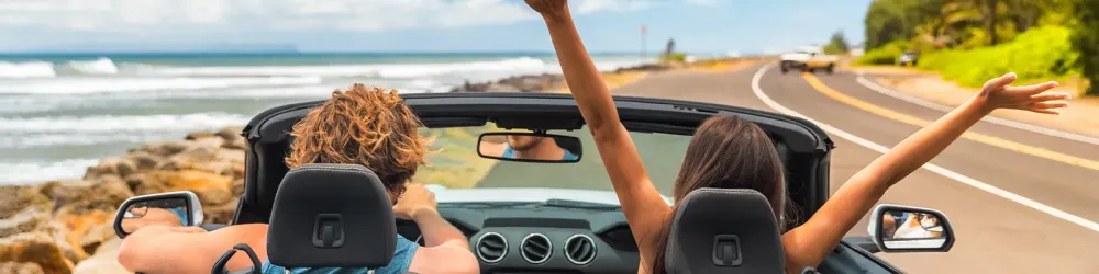 Road trip summer vacation fun young couple driving convertible car on holiday travel destination banner panoramic. Woman with arms up happy, man driver having fun.